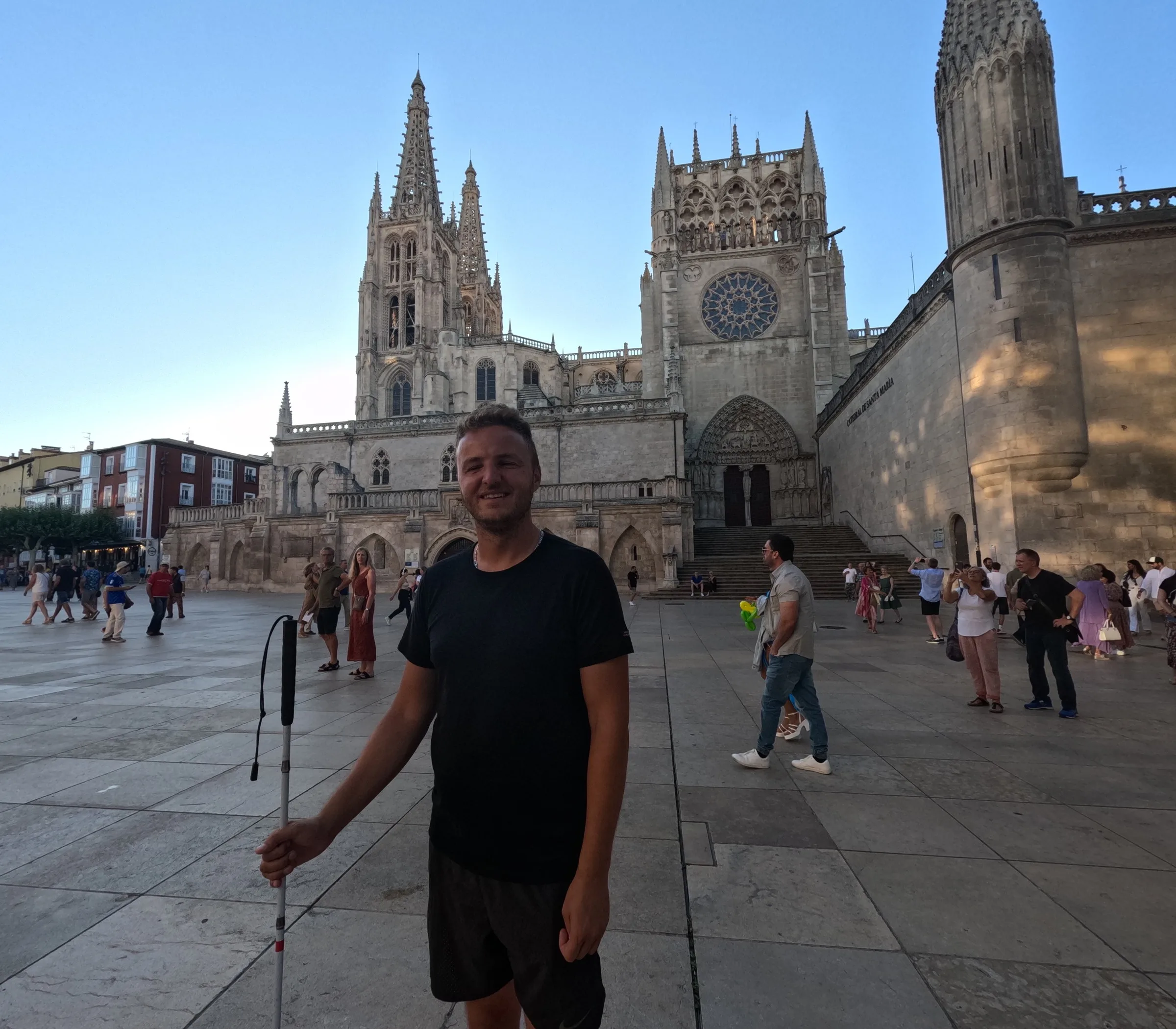 Edis smiler foran Burgos' gotiske katedral, iført sort t-shirt og med sin hvide stok, på en solbeskinnet plads med andre besøgende.