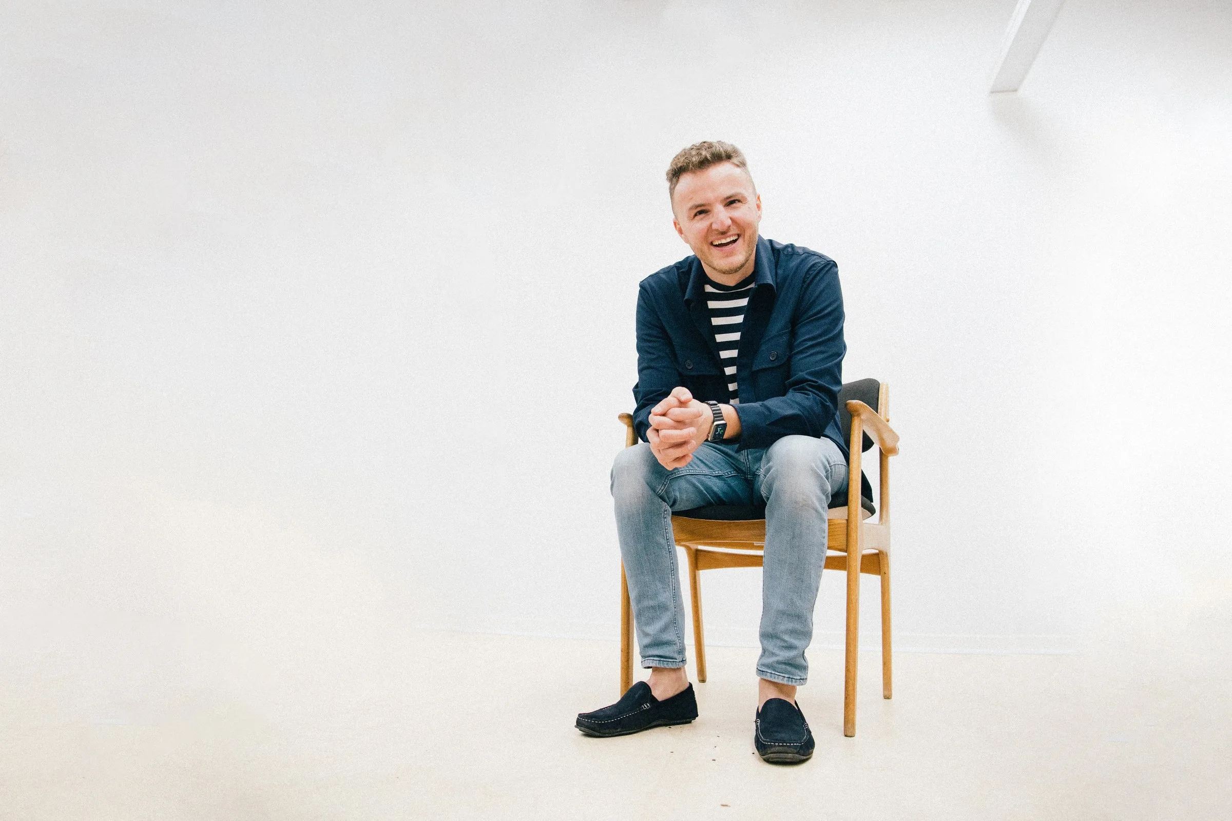 Edis sidder og griner på en træstol i et hvidt studie, iført mørkeblå jakke, stribet trøje og lyse jeans, med hænderne foldet.