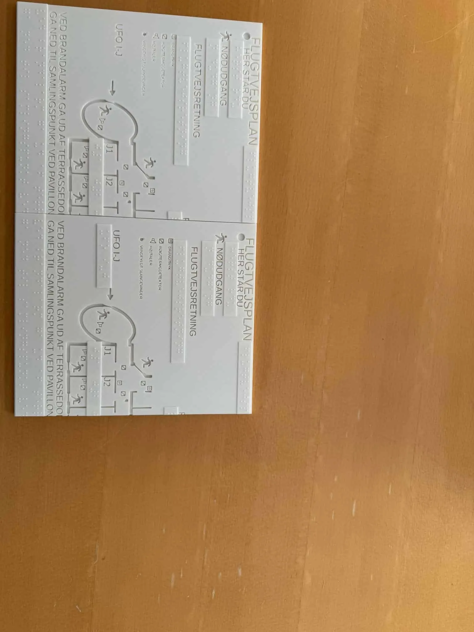 Close-up of a tactile evacuation plan in white plastic with raised corridors, rooms and braille, lying on a light wooden table.